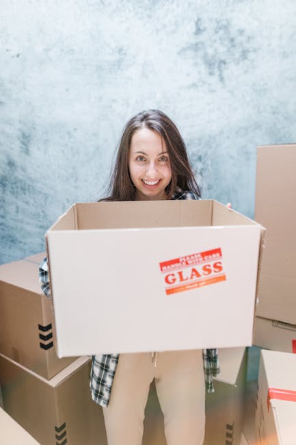 A young woman with long brown hair and a cheerful expression is standing indoors in front of a light-colored, textured wall, holding an empty cardboard moving box with a red 'GLASS' label on one side. Several other packed cardboard boxes of varying sizes, some with black tape or red tape, are arranged around her, indicating an active packing or moving process. She is wearing a plaid shirt over a light-colored top and beige trousers. The environment appears well-lit with natural or bright artificial light, and the scene captures the stages of home packing and furniture transport as part of a professional removals process. The image reflects elements of a home relocation undertaken by Removal Van Knightsbridge, emphasizing packing materials, moving boxes, and the preparation for furniture transport within a house or apartment setting.