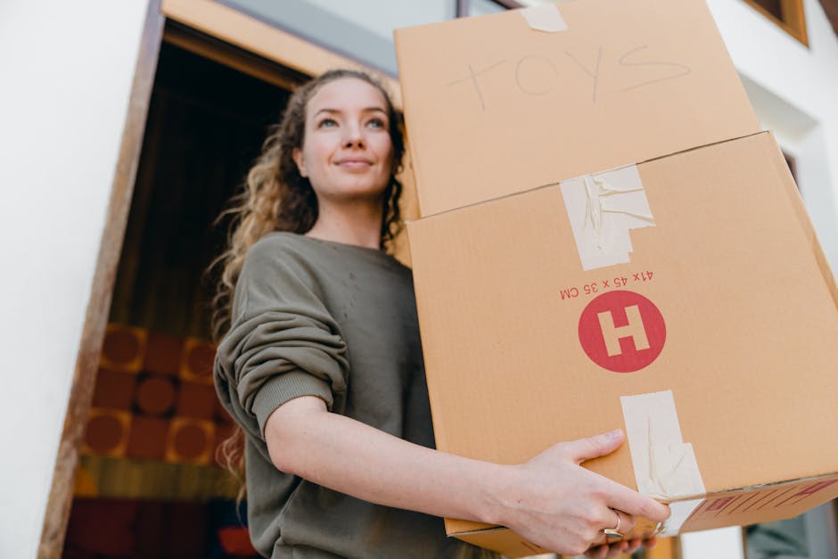 A young woman with curly hair, dressed in a long-sleeved grey top, is standing inside a house near a doorway holding two cardboard boxes stacked one on top of the other. The top box is labelled 'TOYS' handwritten in blue marker, while the bottom box features printed symbols and text indicating packing material, with a white strip of tape securing it. The boxes are made of brown corrugated cardboard, used for home relocation, and include packing tape. The woman is preparing for a move with professional removals, and her expression appears calm as she looks slightly upward. The environment suggests an indoor setting with a wooden staircase and decorative elements in the background, illuminated by soft natural lighting, typical of packing and moving procedures facilitated by Removal Van Knightsbridge.