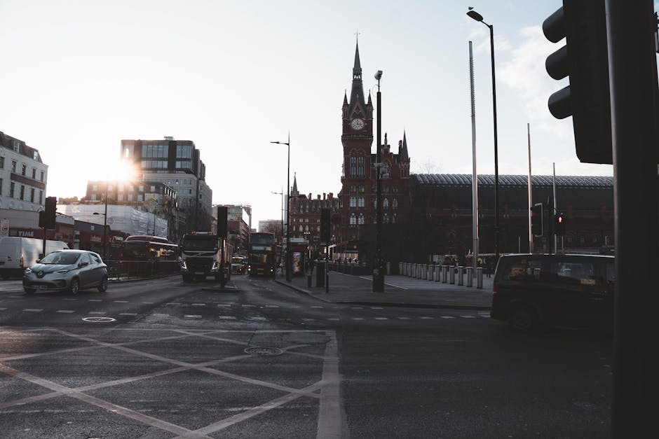 The image shows a busy street scene in Knightsbridge with several vehicles, including a white car, a black van, and multiple buses, positioned at a traffic light intersection. The background features a historic, red-brick building with a tall clock tower and pointed spires, characteristic of the area's architecture. The street is lined with modern and older buildings, some of which are partially shadowed by the setting or rising sun, casting a warm glow behind the vehicles. In the foreground, pavement markings and pedestrian crossing lines are visible, along with traffic lights mounted on black poles. The scene suggests an urban area with active vehicle movement typical of a central location, where professional home relocation services such as those offered by Removal Van Knightsbridge could be involved in coordinating furniture transport and packing during the loading process, especially in areas with potential delays caused by traffic or busy streets.