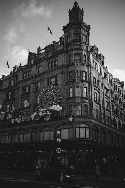A black and white photograph of a multi-story building on Sloane Street in Knightsbridge, SW1X, featuring ornate architectural details and large windows. The building has a prominent vertical sign displaying 'HAR' and additional signage indicating a theatre or entertainment venue, with decorative lighting and signage attached to the façade. The ground level shows a busy street scene with pedestrians and parked vehicles, indicative of a commercial area. In front of the building, a removal van from Removal Van Knightsbridge is parked on the pavement, with part of a trolley and packaging materials such as cardboard boxes and moving blankets visible nearby, suggesting active furniture transport or home relocation processes during the loading or unloading stage of a removals service.