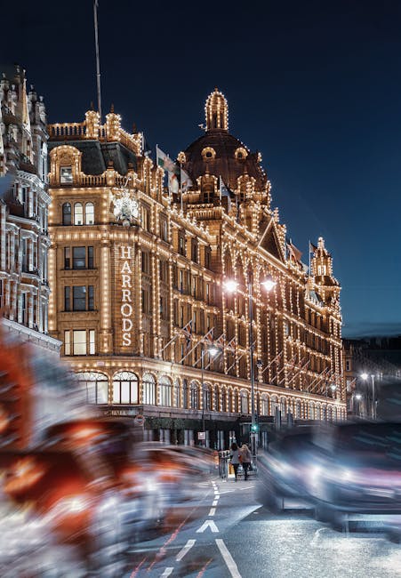A nighttime view of a historic building on Sloane Street in Knightsbridge, illuminated with numerous decorative string lights outlining its architectural features, including domes and turrets. The building, identified by a vertical sign reading 'HARDRODS', is situated on a busy street with some blurred moving vehicles in the foreground, indicating motion and active traffic. The scene is captured from the pavement, and the sky is dark, highlighting the warm glow of the lights against the night sky. This setting reflects an urban environment that could be involved in house removals or moving services, such as those provided by Removal Van Knightsbridge, with preparations implied for logistics and transport of household items, although no furniture or moving activity is visible in the image.