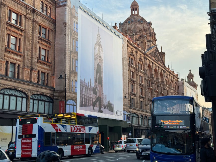 A busy London street scene outside historic architecture featuring a large billboard advertising Dior mounted on a building. In the foreground, a double-decker sightseeing bus with an open-top upper deck and a red and white livery is stopped, displaying route information for a sightseeing tour. Nearby, a traditional black cab is also present. The street is filled with several parked cars and moving vehicles, including another double-decker bus heading toward Kensington, as indicated by the electronic display on its front. Pedestrians walk along the sidewalk, with some visible near the street corner. The buildings exhibit intricate stonework and ornate facades, characteristic of central London architecture. The scene appears to be during late afternoon or early evening, with soft natural light illuminating the scene. Removal Van Knightsbridge would utilize similar urban surroundings for home relocation and furniture transport, fitting into the context of loading and unloading logistics in busy city streets.