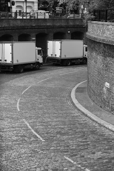 A black-and-white photograph showing a cobbled street with a curved layout, partially enclosed by a brick wall on the right side. In the background, there are two white removal vans parked parallel to each other, positioned near an underpass with arches. The vans are part of a house relocation fleet used by Removal Van Knightsbridge, likely engaged in furniture transport or packing and moving activities. The vans are situated close to the building's entrance, which is adjacent to a pavement with a railing, suggesting a nearby residential or commercial property. The loading area appears spacious enough for the vans to facilitate the loading and unloading of boxes, appliances, and furniture during a house move. The lighting is natural, with shadows indicating that the image was taken during daylight hours, emphasizing the logistics involved in a professional removals service in London.