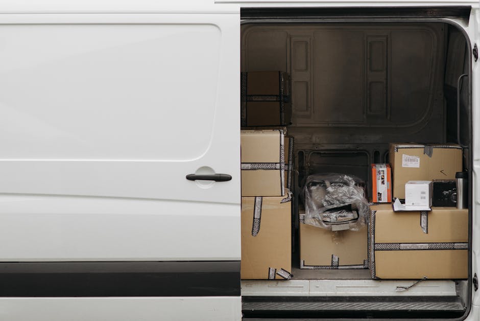 Inside the loading area of a white removal van, four large cardboard boxes with white labels are neatly stacked, three on the floor and one placed on top. The boxes are secured with plastic wrap and a