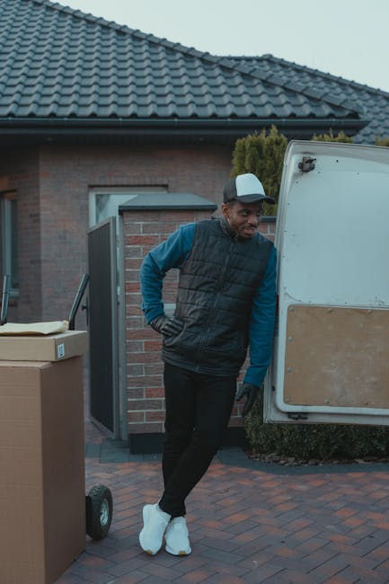 A man wearing a black padded vest, blue long-sleeve shirt, black trousers, white sneakers, and a grey cap stands outside a residential property, leaning against an open vehicle door. The scene takes p