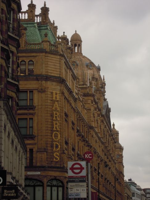 A busy street scene in Knightsbridge, London, with rows of historic multi-storey buildings featuring detailed stone facades, large windows, and decorative elements. The street is populated with parked