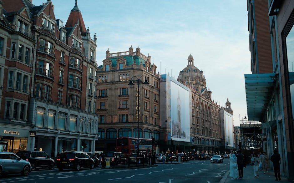 A street scene near Harrods on Brompton Road in Knightsbridge, London, showing a row of historic multi-storey buildings with architectural details such as decorative cornices and turrets. Parked cars line the curb, while pedestrians walk along the sidewalk, some dressed in casual clothing. On the right, a modern building with a glass canopy extends over the sidewalk. The street is bustling, with a red double-decker bus visible in the background. The sky is clear with a few clouds, and the natural daylight highlights the intricate facades of the buildings, creating a lively urban atmosphere typical of the Knightsbridge area. This setting suggests a vibrant location where moving services like those provided by Removal Van Knightsbridge could operate, especially during home relocations or furniture transport activities.