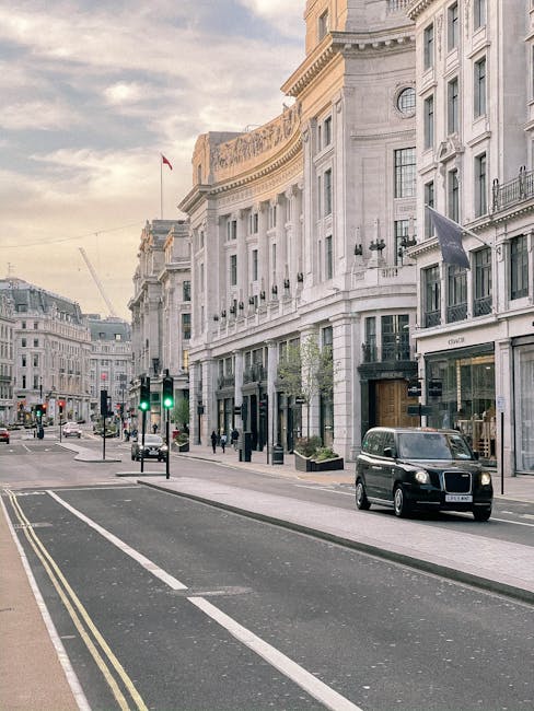 A street scene in Knightsbridge with elegant white stone buildings featuring large windows and decorative architectural details, lined along the pavement. A black van from Removal Van Knightsbridge is parked on the right side of the street, adjacent to the entrance of a high-end retail store with a black awning. Several cardboard boxes and wrapped furniture are visible inside the open rear of the van, indicating an active home relocation or furniture transport process. The street has a few pedestrians walking along the sidewalk and traffic lights showing green, with a bicycle lane marked on the road. The overall scene is illuminated by soft natural light, suggesting early morning or late afternoon, reflecting a typical urban environment engaged in packing and moving activities coordinated by [COMPANY_NAME], supporting efficient house removals near Harrods Brompton Road Knightsbridge.
