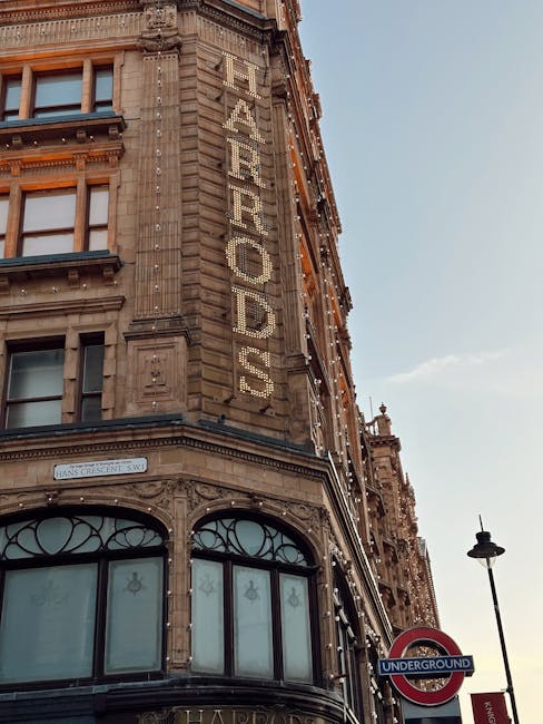 A street scene near Harrods on Brompton Road in Knightsbridge, London, showing a row of historic multi-storey buildings with architectural details such as decorative cornices and turrets. Parked cars line the curb, while pedestrians walk along the sidewalk, some dressed in casual clothing. On the right, a modern building with a glass canopy extends over the sidewalk. The street is bustling, with a red double-decker bus visible in the background. The sky is clear with a few clouds, and the natural daylight highlights the intricate facades of the buildings, creating a lively urban atmosphere typical of the Knightsbridge area. This setting suggests a vibrant location where moving services like those provided by Removal Van Knightsbridge could operate, especially during home relocations or furniture transport activities.