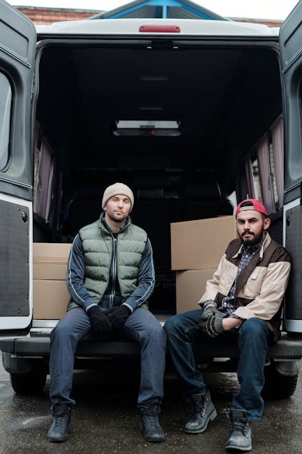 Two men are seated on the open back doors of a large grey removal van, which is parked outdoors during daylight hours. The van's interior is visible behind them, with multiple cardboard boxes of varying sizes secured inside, some stacked on top of each other. One man wears a beige beanie, a green puffer vest over a navy blue jacket, and dark trousers, while the other is dressed in a red cap worn backwards, a beige and black jacket, and dark pants. Both men have gloves on and appear to be taking a break during the loading process of furniture and packing materials typically involved in house removals, with the exterior environment showing a paved surface and a glimpse of brickwork in the background. This scenario reflects a professional moving or packing operation, consistent with the services offered by Removal Van Knightsbridge for home relocation tasks involving tight access jobs and careful furniture transport.