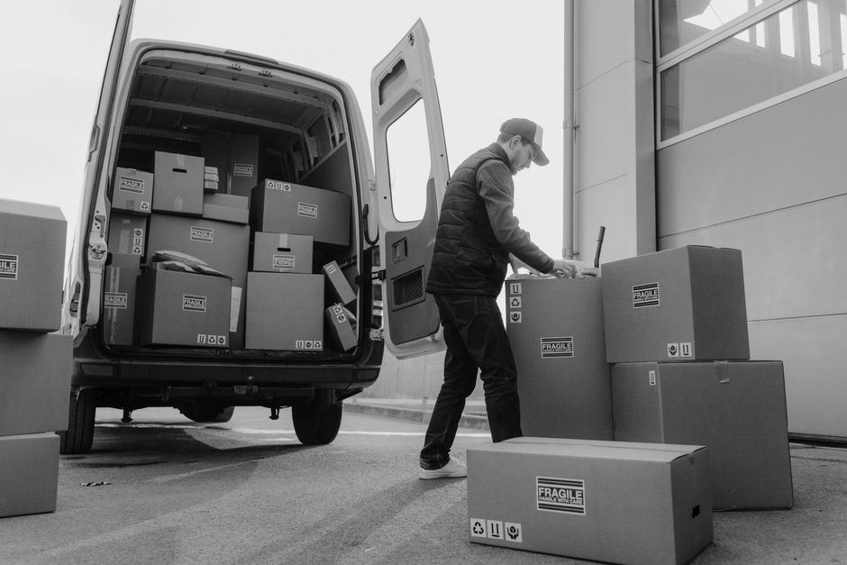 A man wearing a cap and outdoor clothing is engaged in packing or unloading cardboard boxes labeled 'fragile' during a home relocation process outside a building with a large roller shutter door. He is standing beside a white moving van with its back doors open, revealing numerous packed boxes of varying sizes inside, some stacked on top of each other. The man is placing or preparing additional boxes, which are arranged on the pavement next to the van, some on the ground and others on a trolley, for loading or unloading. The boxes are secured with plastic wrap and cardboard with printed handling symbols. The scene is illuminated by natural light, and the environment appears organized for the loading process of furniture transport or packing and moving services, reflecting the logistics involved in house removals. Removal Van Knightsbridge specializes in such relocation logistics, including managing tight access and ensuring safe, efficient movement of household items.