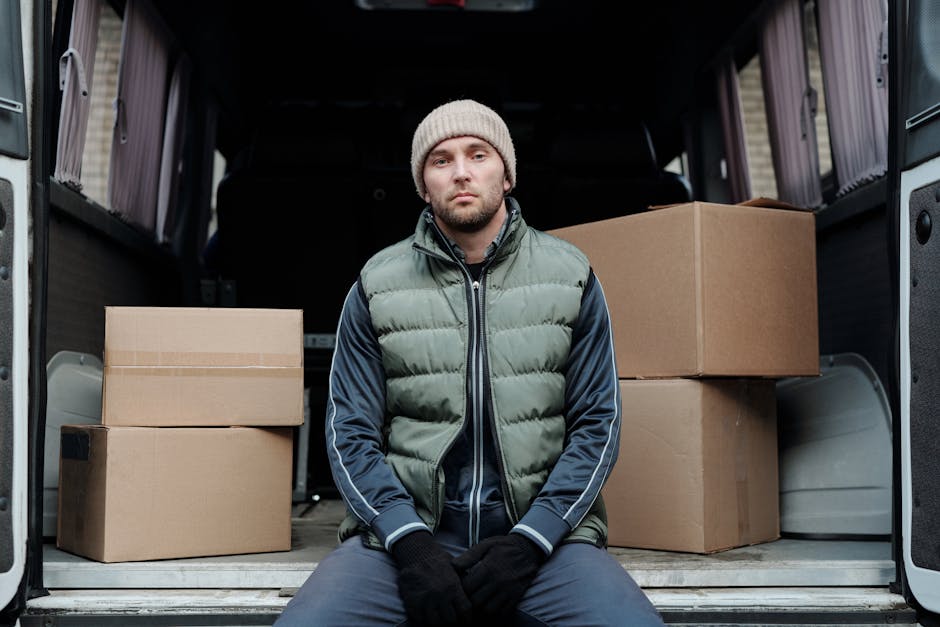 Two men are seated on the open back doors of a large grey removal van, which is parked outdoors during daylight hours. The van's interior is visible behind them, with multiple cardboard boxes of varying sizes secured inside, some stacked on top of each other. One man wears a beige beanie, a green puffer vest over a navy blue jacket, and dark trousers, while the other is dressed in a red cap worn backwards, a beige and black jacket, and dark pants. Both men have gloves on and appear to be taking a break during the loading process of furniture and packing materials typically involved in house removals, with the exterior environment showing a paved surface and a glimpse of brickwork in the background. This scenario reflects a professional moving or packing operation, consistent with the services offered by Removal Van Knightsbridge for home relocation tasks involving tight access jobs and careful furniture transport.