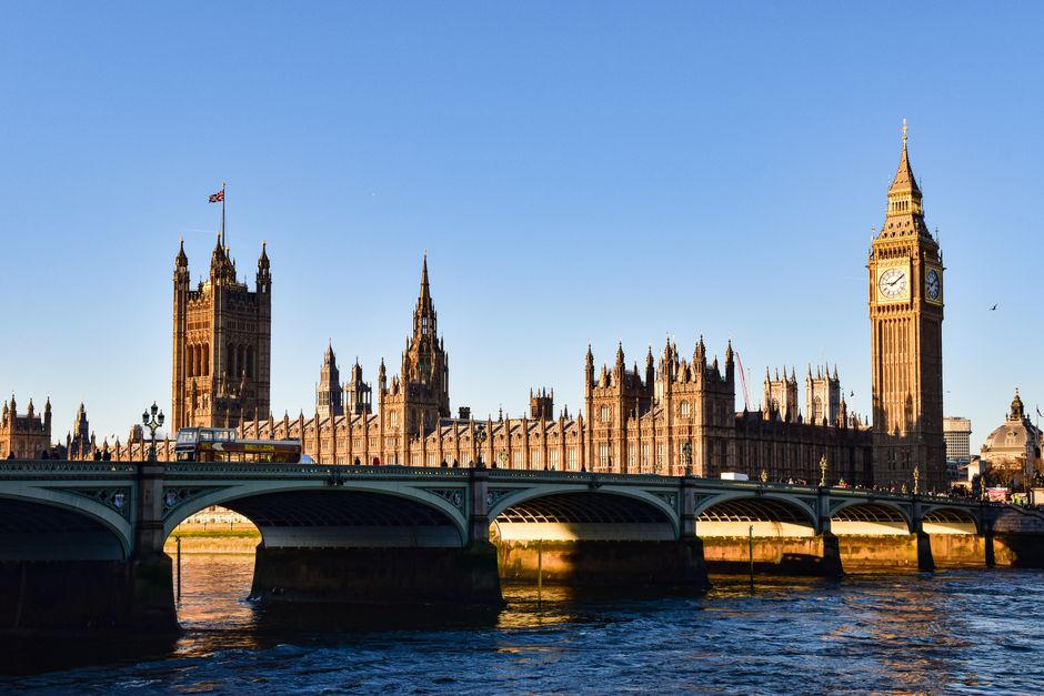 A wide-angle view of the Palace of Westminster in London, featuring the William Ewart Gladstone Memorial Bridge in the foreground with car traffic on it. The iconic clock tower, commonly known as Big Ben, is visible on the right side of the image, showcasing its detailed gothic architecture with a clock face displaying the time. The historic building complex is illuminated by natural light, with clear blue sky overhead. The river Thames flows beneath the bridge, reflecting the structure and the sunlight. This scene is related to London’s central area where house removals and furniture transport may occur, and it highlights the importance of obtaining Westminster council permits for such relocation services. As performed by Removal Van Knightsbridge, moving logistics in this area often require careful planning around local regulations and historical environment considerations seen here.