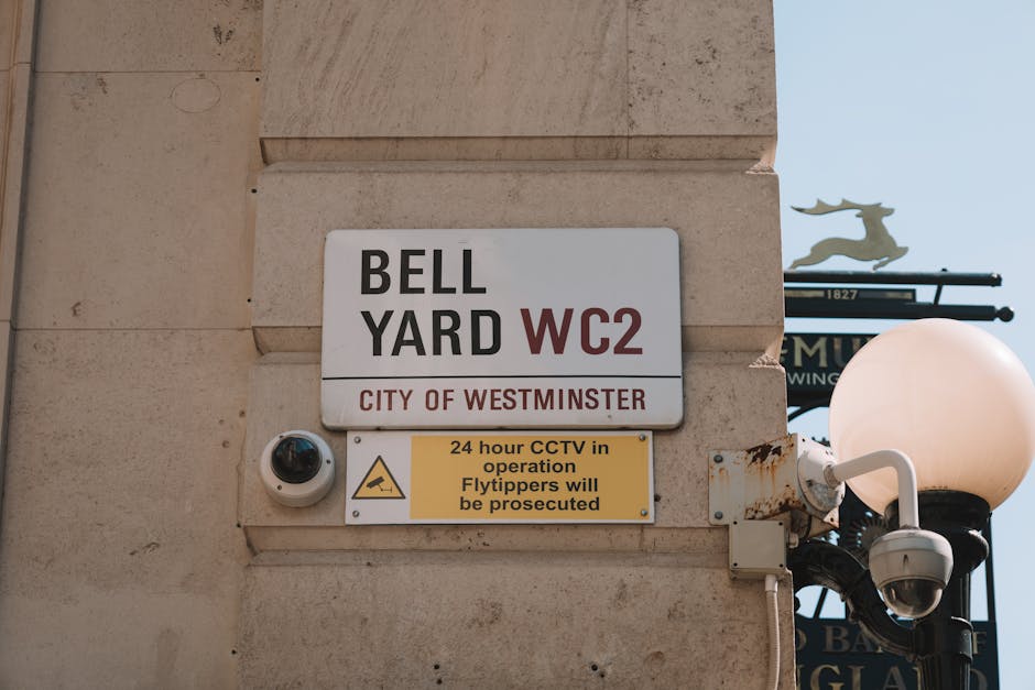 The image shows a close-up of a building corner with a white sign indicating 'BELL YARD WC2' in bold black and red lettering, and 'CITY OF WESTMINSTER' beneath it. Below the sign, there is a smaller yellow warning sign stating '24 hour CCTV in operation Flytippers will be prosecuted,' accompanied by a CCTV camera mounted on the wall. To the right, a traditional black lamp post with a round, white-globed light is partially visible, along with a black street sign featuring a silhouette of a reindeer attached to a decorative metal arm. The scene occurs outdoors during daytime with natural lighting and a clear sky in the background. The image depicts a typical urban area relevant to house removals and moving logistics, where parking and security considerations such as CCTV are important for efficient home relocation and furniture transport under Westminster council permit rules, as referenced on the page about removals at removalvanknightsbridge.co.uk.