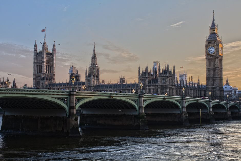 A wide-angle view of the Palace of Westminster in London, featuring the William Ewart Gladstone Memorial Bridge in the foreground with car traffic on it. The iconic clock tower, commonly known as Big Ben, is visible on the right side of the image, showcasing its detailed gothic architecture with a clock face displaying the time. The historic building complex is illuminated by natural light, with clear blue sky overhead. The river Thames flows beneath the bridge, reflecting the structure and the sunlight. This scene is related to London’s central area where house removals and furniture transport may occur, and it highlights the importance of obtaining Westminster council permits for such relocation services. As performed by Removal Van Knightsbridge, moving logistics in this area often require careful planning around local regulations and historical environment considerations seen here.