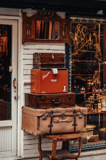 A collection of vintage and antique objects arranged outside a house in a relocation setting. The display includes a large, light brown leather suitcase placed on a small wooden table, with a smaller, dark brown wooden chest stacked on top. Above the chest is a red vintage petrol canister with a white logo and a metal handle, and next to it is a smaller wooden box with a metal latch. To the left, part of a glass door with a white frame is visible, while to the right, a window display shows various brass and metallic items, possibly antique household objects or collectibles, with some decorative elements. The background features a white wooden wall beneath a dark roof, with a small wooden framed mirror or picture hanging above the objects. The scene suggests a setting aligned with house removals, packing, and moving logistics, highlighting the careful handling of fragile antiques during a home relocation, in an outdoor space adjacent to the property.